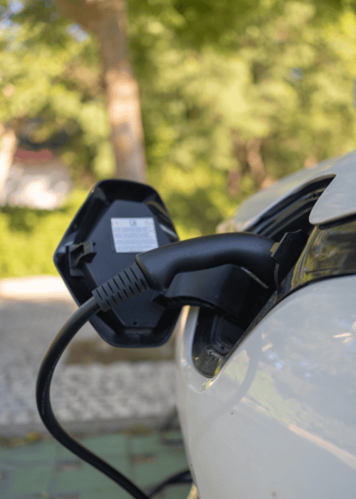 A Close-Up Of An Ev Car Charger Plugged Into An Electric Vehicle’s Port, With Trees And Pavement Softly Blurred In The Background.
